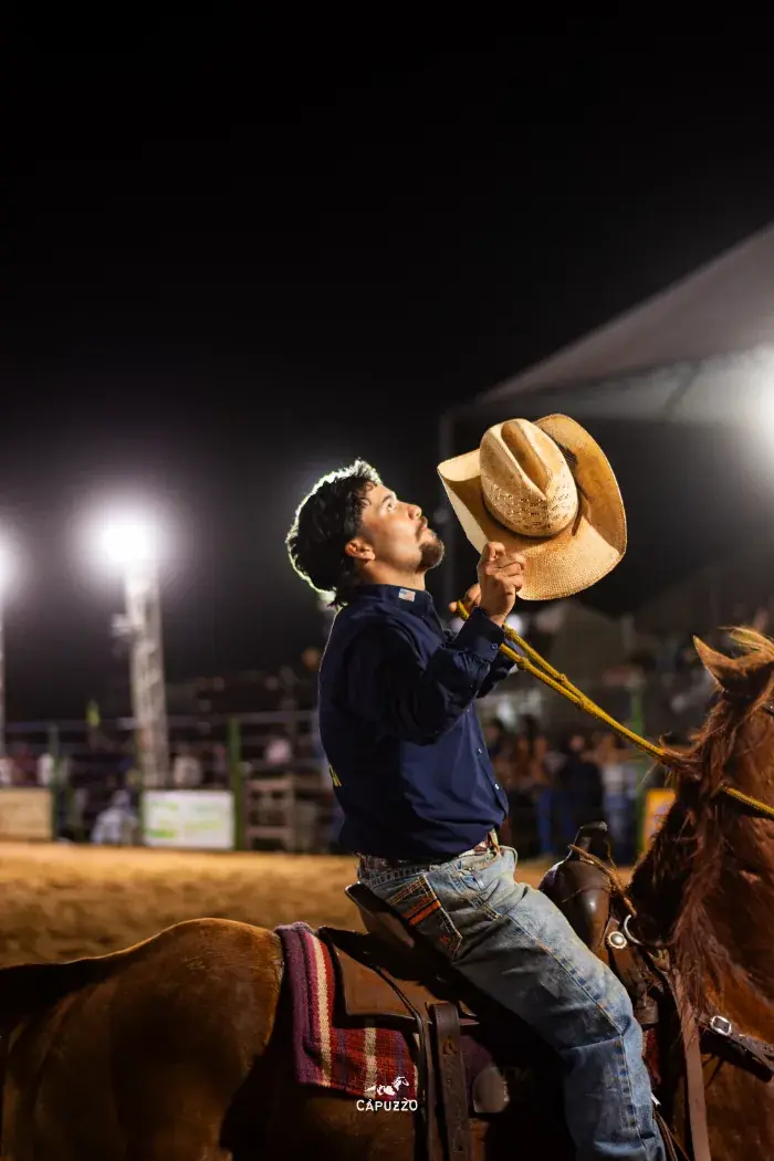 Peão a cavalo levantando o chapéu sob a luz da arena à noite
